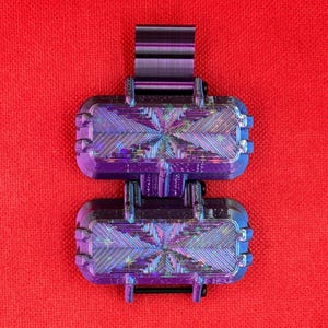 Puede incluir: Un contenedor morado de dos compartimentos impreso en 3D, con un dise&ntilde;o articulado y un clip superior. Las unidades rectangulares tienen esquinas redondeadas y una superficie texturizada e iridiscente que refleja la luz con un efecto estelar.