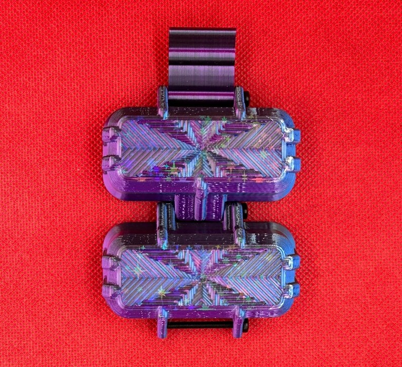 Puede incluir: Un contenedor morado de dos compartimentos impreso en 3D, con un dise&ntilde;o articulado y un clip superior. Las unidades rectangulares tienen esquinas redondeadas y una superficie texturizada e iridiscente que refleja la luz con un efecto estelar.