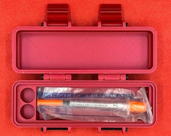 May include: A rectangular magenta plastic storage case with two hinged latches. The open case contains a single-use U-100 insulin syringe in a clear plastic wrapper, positioned next to two circular cutouts in the molded interior base.