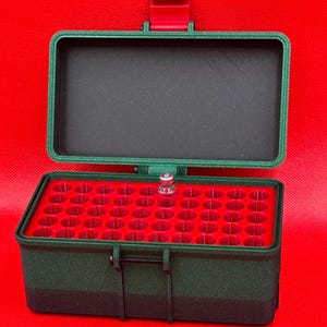 May include: A dark green rectangular storage case with a hinged lid and a red latch. The interior features a red insert with multiple circular slots for small items, and one small clear glass vial is placed in one of the slots.