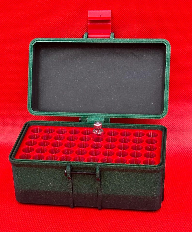 May include: A dark green rectangular storage case with a hinged lid and a red latch. The interior features a red insert with multiple circular slots for small items, and one small clear glass vial is placed in one of the slots.