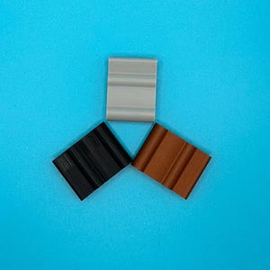 May include: Three rectangular clips arranged in a triangular formation. One clip is light gray, one is solid black, and one is a metallic copper color. Each clip features a distinct grooved surface with two parallel vertical channels.