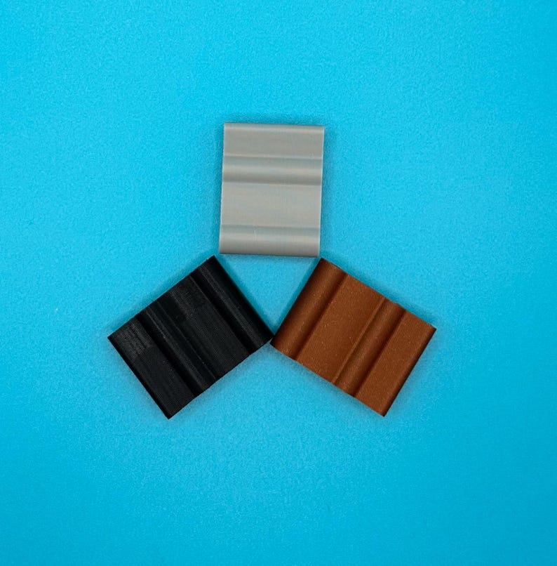 May include: Three rectangular clips arranged in a triangular formation. One clip is light gray, one is solid black, and one is a metallic copper color. Each clip features a distinct grooved surface with two parallel vertical channels.