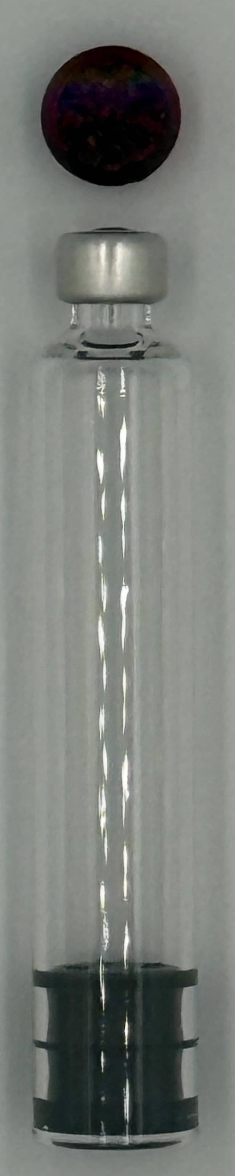 May include: A clear glass vial with a silver metal crimp cap and a dark, iridescent circular topper positioned above it. The vial features a cylindrical body and a black rubber stopper visible at the base, creating a distinct contrast.