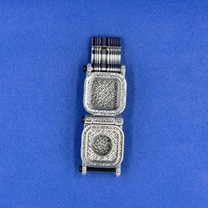 K&ouml;nnte beinhalten: Ein silberfarbenes Uhrengeh&auml;use aus Metall mit zwei quadratischen F&auml;chern und komplexen, strukturierten Mustern im Inneren. Das obere Fach ist leer, w&auml;hrend das untere Fach eine kreisf&ouml;rmige Vertiefung aufweist. Ein gestreiftes Armband ist befestigt.