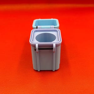 May include: A small, square, light blue container with a hinged lid that is open. The container features a circular opening on the top surface and a thin black metal pin acting as a hinge mechanism on the side of the box structure.
