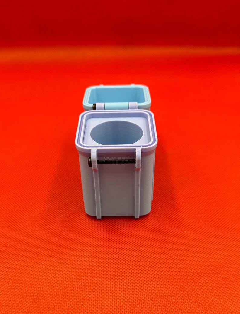 May include: A small, square, light blue container with a hinged lid that is open. The container features a circular opening on the top surface and a thin black metal pin acting as a hinge mechanism on the side of the box structure.