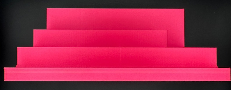May include: A bright pink tiered display stand featuring three distinct levels of varying heights. The structure is composed of horizontal rectangular platforms arranged in a stepped formation, providing a clear surface for organizing items.