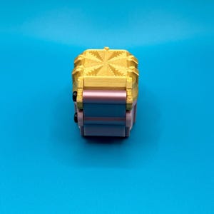 K&ouml;nnte beinhalten: Ein kleines, dreidimensionales Objekt mit einem goldfarbenen Oberteil, das ein strukturiertes, sternartiges geometrisches Muster aufweist. Die Basis besteht aus gestapelten, glatten, hellrosa und blauen rechteckigen Komponenten mit zwei kleinen schwarzen runden Akzenten.