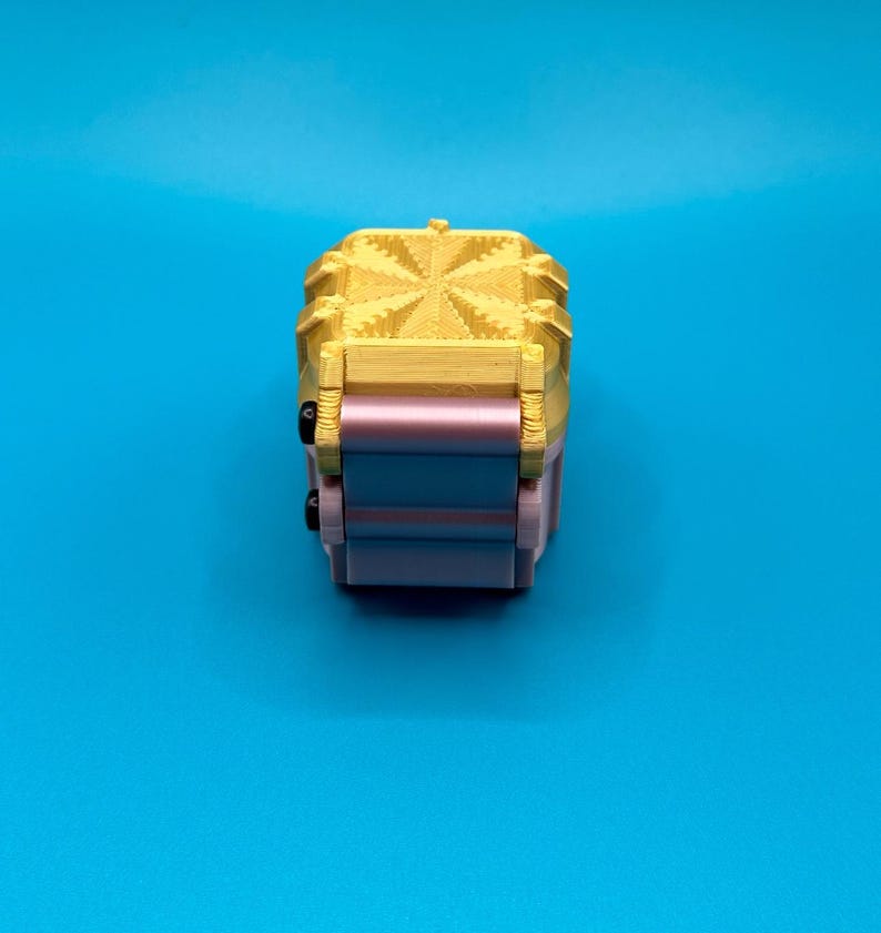 K&ouml;nnte beinhalten: Ein kleines, dreidimensionales Objekt mit einem goldfarbenen Oberteil, das ein strukturiertes, sternartiges geometrisches Muster aufweist. Die Basis besteht aus gestapelten, glatten, hellrosa und blauen rechteckigen Komponenten mit zwei kleinen schwarzen runden Akzenten.