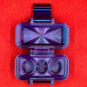 Puede incluir: Un estuche de almacenamiento rectangular con bisagra y acabado iridiscente en tonos morado y azul met&aacute;lico. El interior presenta un patr&oacute;n geom&eacute;trico texturizado con tres hendiduras circulares en la base y una secci&oacute;n superior hueca.