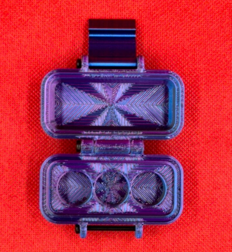 Puede incluir: Un estuche de almacenamiento rectangular con bisagra y acabado iridiscente en tonos morado y azul met&aacute;lico. El interior presenta un patr&oacute;n geom&eacute;trico texturizado con tres hendiduras circulares en la base y una secci&oacute;n superior hueca.