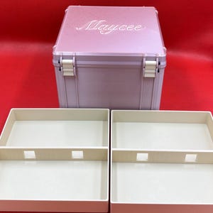 以下が含まれることがあります： 蓋にMayceeの名前が刻印されたライトピンクの正方形の収納ボックス。白いヒンジが付いており、中央に仕切りと四角い切り抜きがあるクリーム色の長方形のトレイ2つが手前に配置されています。