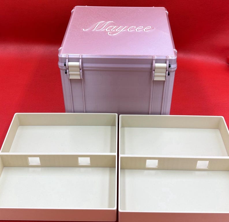 以下が含まれることがあります： 蓋にMayceeの名前が刻印されたライトピンクの正方形の収納ボックス。白いヒンジが付いており、中央に仕切りと四角い切り抜きがあるクリーム色の長方形のトレイ2つが手前に配置されています。