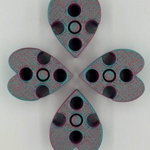 May include: Four heart-shaped objects arranged in a cloverleaf pattern. Each object has a speckled teal and pink texture with black circular indentations. The objects appear to be made of a 3D-printed material.