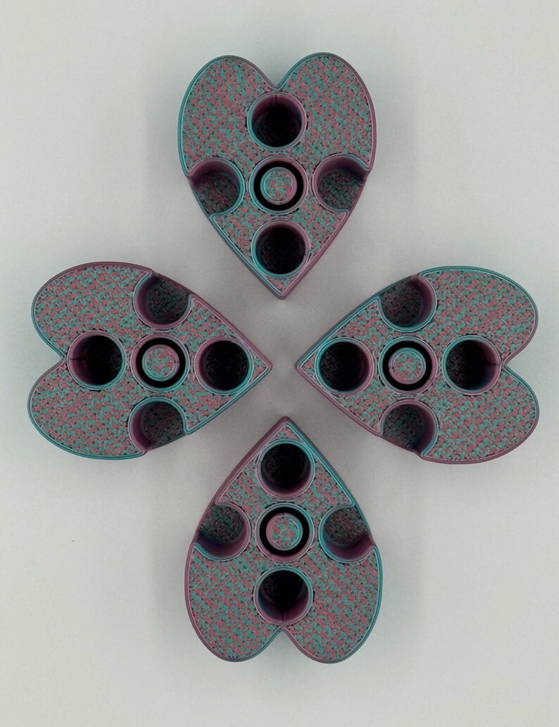 May include: Four heart-shaped objects arranged in a cloverleaf pattern. Each object has a speckled teal and pink texture with black circular indentations. The objects appear to be made of a 3D-printed material.
