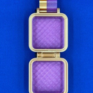 May include: A square, hinged container featuring a gold-colored frame and a translucent, glittery purple interior. The object has a vertical gold and purple tab at the top and a textured, layered surface finish consistent with 3D printing.
