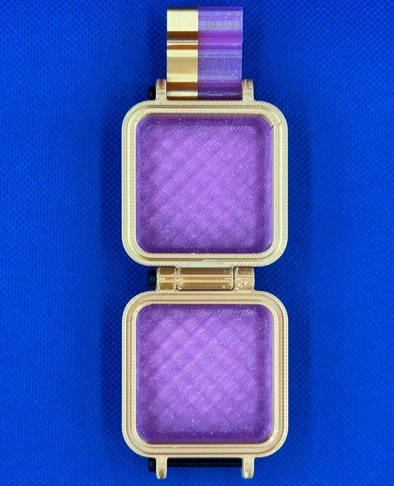 May include: A square, hinged container featuring a gold-colored frame and a translucent, glittery purple interior. The object has a vertical gold and purple tab at the top and a textured, layered surface finish consistent with 3D printing.
