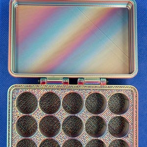 May include: A rectangular storage case with a hinged lid, featuring a multi-colored iridescent finish. The interior base contains fifteen circular compartments arranged in three rows of five, while the lid interior is smooth and flat.