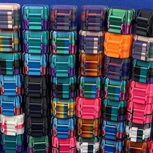 May include: A collection of small, rectangular, articulated fidget toys arranged in vertical stacks. Each toy features a multi-segmented design with various color combinations, including solid black, bright orange, pink, white, and metallic gradients.