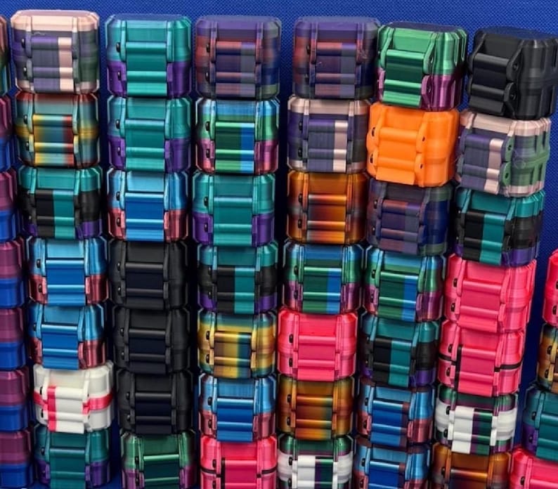 May include: A collection of small, rectangular, articulated fidget toys arranged in vertical stacks. Each toy features a multi-segmented design with various color combinations, including solid black, bright orange, pink, white, and metallic gradients.