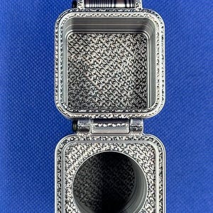 May include: A silver-toned, rectangular, hinged metal container with a textured, repeating geometric pattern on its interior surfaces. The top section is an open square compartment, while the bottom section features a circular opening.