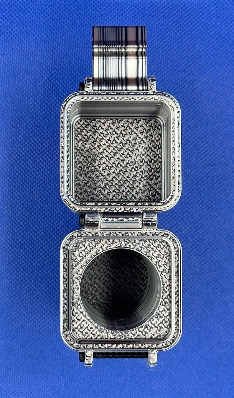 May include: A silver-toned, rectangular, hinged metal container with a textured, repeating geometric pattern on its interior surfaces. The top section is an open square compartment, while the bottom section features a circular opening.