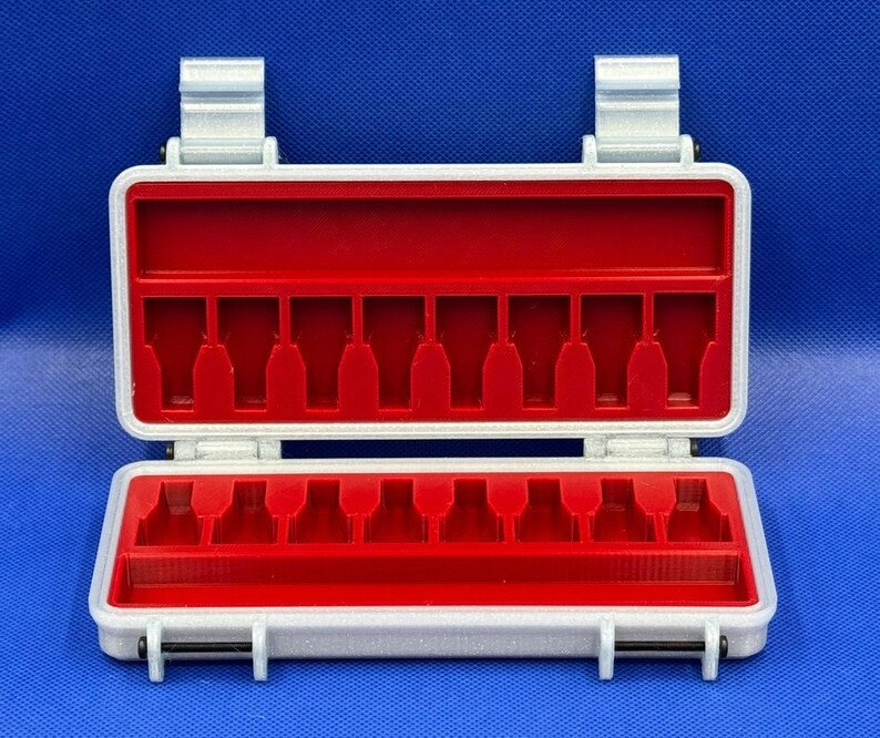 May include: A rectangular silver-colored storage case with a hinged lid and two latches. The interior features a bright red insert with ten vertical slots designed for holding small items, plus a long horizontal compartment at the base.