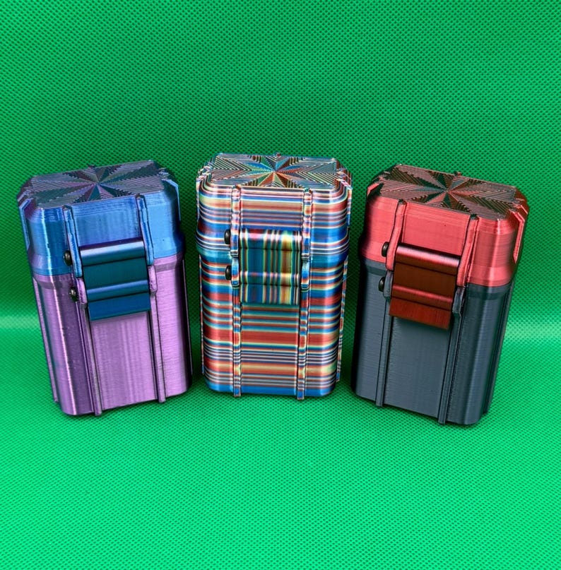 Puede incluir: Tres cajas de almacenamiento rectangulares con tapas abatibles y cierres frontales. La caja de la izquierda es azul y morada, la central presenta un patr&oacute;n de rayas horizontales multicolores y la de la derecha es roja y gris oscura.