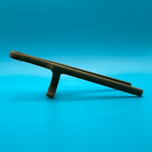 May include: A matte brown, elongated, slender object with a central support strut, resembling a stylized stand or holder. The item features a smooth, uniform surface texture and a horizontal orientation, resting on its single vertical base.
