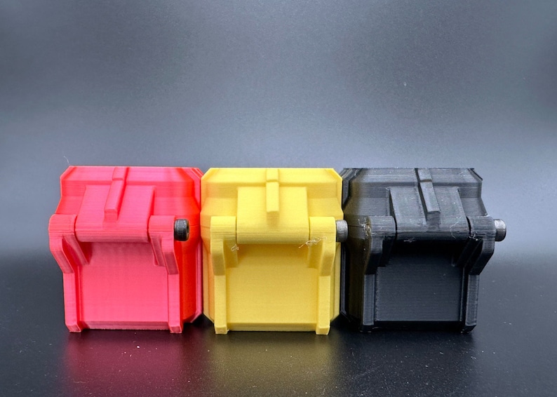 K&ouml;nnte beinhalten: Drei kleine, rechteckige, 3D-gedruckte Beh&auml;lter mit Klappdeckeln, die nebeneinander angeordnet sind. Die Beh&auml;lter sind rot, gelb und schwarz und verf&uuml;gen jeweils &uuml;ber einen zentralen vertikalen Steg und ein kleines Metallstift-Scharnier an der Seite.