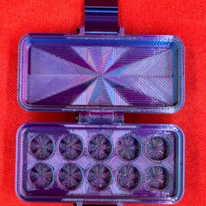 May include: A rectangular, hinged storage case with a metallic purple and blue gradient finish. The interior lid features a radiating geometric pattern, while the base contains ten circular compartments, each with a textured snowflake design.
