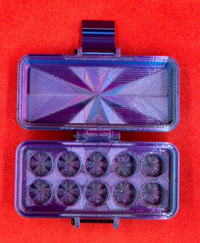 May include: A rectangular, hinged storage case with a metallic purple and blue gradient finish. The interior lid features a radiating geometric pattern, while the base contains ten circular compartments, each with a textured snowflake design.