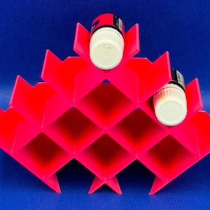 Puede incluir: Un estante de almacenamiento de pl&aacute;stico rosa ne&oacute;n brillante con un patr&oacute;n de rejilla geom&eacute;trico en forma de diamante, dise&ntilde;ado para sostener botellas cil&iacute;ndricas peque&ntilde;as. Dos frascos peque&ntilde;os con tapas blancas est&aacute;n insertados.