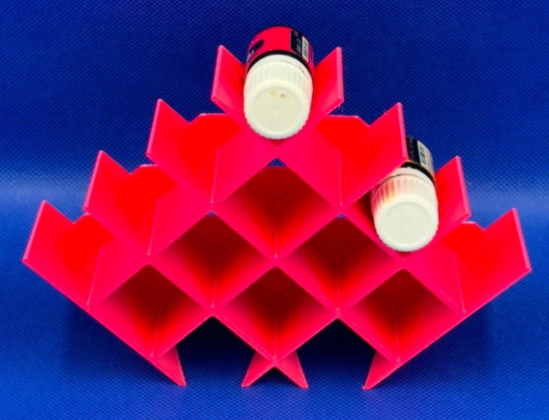 Puede incluir: Un estante de almacenamiento de pl&aacute;stico rosa ne&oacute;n brillante con un patr&oacute;n de rejilla geom&eacute;trico en forma de diamante, dise&ntilde;ado para sostener botellas cil&iacute;ndricas peque&ntilde;as. Dos frascos peque&ntilde;os con tapas blancas est&aacute;n insertados.