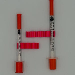 May include: Two insulin syringes with orange caps and plungers, alongside two bright pink syringe holders. The syringes are clear with black measurement markings. The holders have a ridged design.