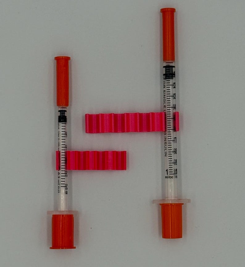 May include: Two insulin syringes with orange caps and plungers, alongside two bright pink syringe holders. The syringes are clear with black measurement markings. The holders have a ridged design.