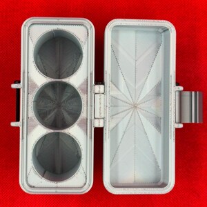 May include: A light gray, rectangular, 3D-printed storage case with a hinged lid. The left side features three circular compartments, while the right side contains a single, open rectangular compartment with a starburst pattern.