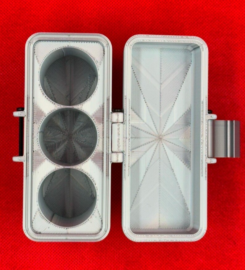 May include: A light gray, rectangular, 3D-printed storage case with a hinged lid. The left side features three circular compartments, while the right side contains a single, open rectangular compartment with a starburst pattern.