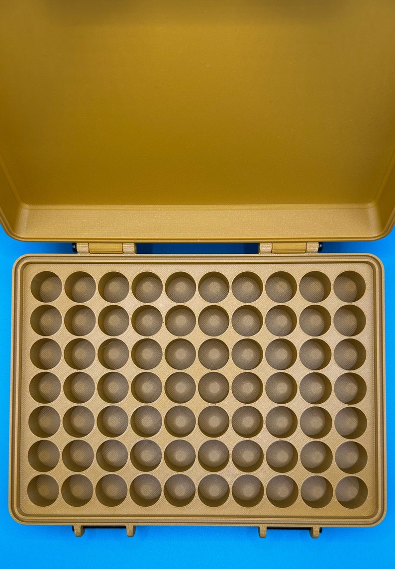 May include: A gold-colored storage case featuring an open lid and a base with a grid of sixty circular indentations. The interior is structured with a uniform pattern of holes designed for holding small items, all contained within a rectangular frame.