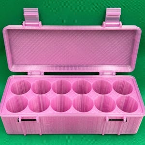 May include: A light pink rectangular storage case with a hinged lid and two secure latches. The interior features twelve circular compartments arranged in two rows of six, designed for holding small cylindrical items securely.