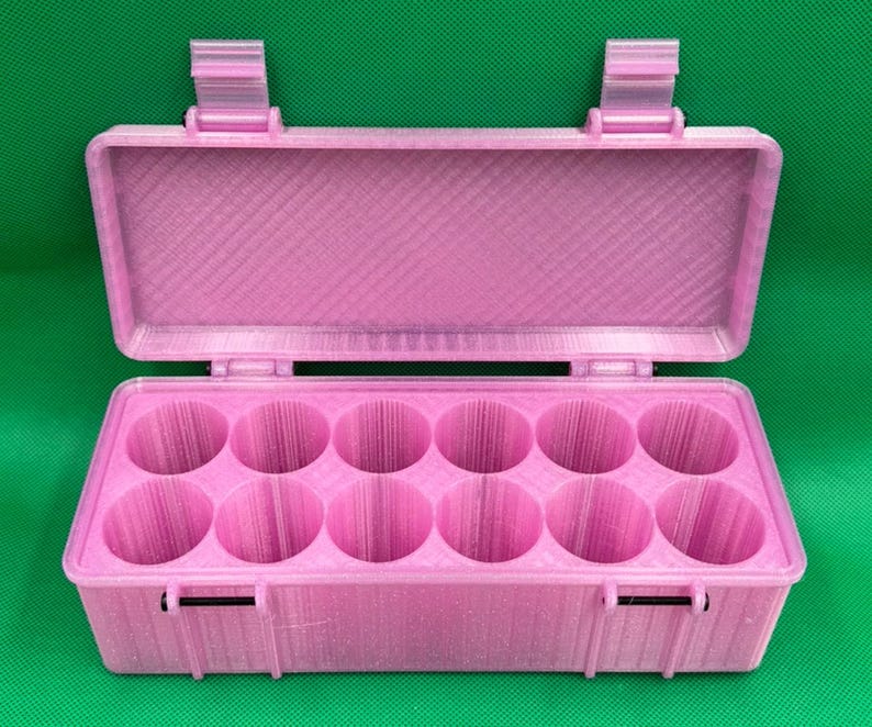 May include: A light pink rectangular storage case with a hinged lid and two secure latches. The interior features twelve circular compartments arranged in two rows of six, designed for holding small cylindrical items securely.