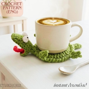 May include: A green crocheted alligator coaster with a red tongue holds a white mug of coffee with latte art. The coaster is handmade and sits on a white surface. A white spoon is next to the coaster.