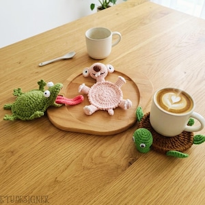 May include: A wooden table displays three crocheted animal coasters: a green frog with a pink tongue, a pink koala, and a brown turtle. A white mug and a cup of coffee with latte art are also present.