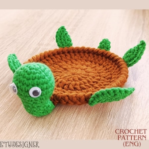 Pode incluir: Uma tartaruga de crochê com casco marrom e cabeça e barbatanas verdes. A tartaruga tem olhos arregalados e está sobre uma superfície de madeira. O texto "CROCHET PATTERN (ENG)" é visível na imagem.