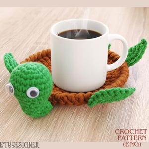 Könnte beinhalten: Eine weiße Kaffeetasse steht auf einem handgefertigten Schildkröten-Untersetzer aus Häkelarbeit. Der Untersetzer ist braun mit grünen flossenartigen Akzenten und einem grünen Schildkrötenkopf mit Wackelaugen.