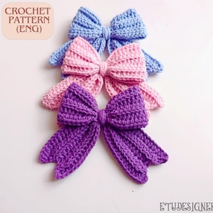 May include: Three handmade crochet bows in shades of blue, pink, and purple. The bows are arranged in a row, with the purple bow at the bottom. The text "CROCHET PATTERN (ENG)" is in the upper left corner.