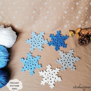 May include: Five crocheted snowflakes in various shades of blue and white. The snowflakes are arranged on a brown surface with the text "CROCHET PATTERN (ENG)" and "etudesigner" visible.