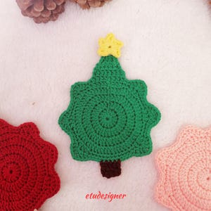Puede incluir: Un posavasos de ganchillo verde con forma de árbol de Navidad con una estrella amarilla en la parte superior y un tronco marrón. También se ven otros dos posavasos de ganchillo, uno rojo y otro rosa. La palabra "etudesigner" está impresa en la parte inferior.