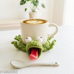 Könnte beinhalten: Eine Keramiktasse mit einem Latte-Art-Design in Herzform steht auf einem grünen gehäkelten Frosch-Untersetzer. Der Frosch hat weiße Augen und eine rosa Zunge. Ein weißer Keramik-Löffel liegt auf dem Tisch. Das Wort "ETUDESIGNER" ist sichtbar.